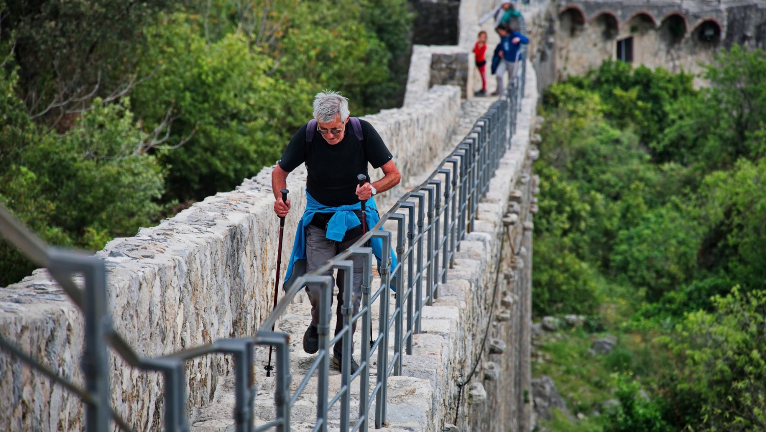 A senior male traveler using walking poles, crossing a bridge